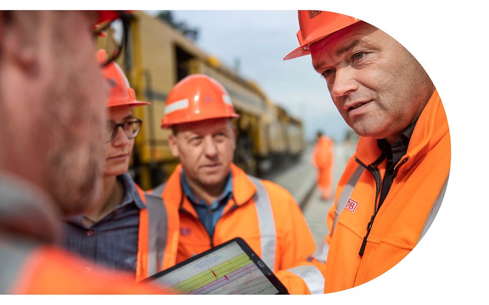 Vier Personen stehen in orangefarbener Schutzkleidung neben einer Gleisbaumaschine und besprechen den Plan auf einem Tablet. 