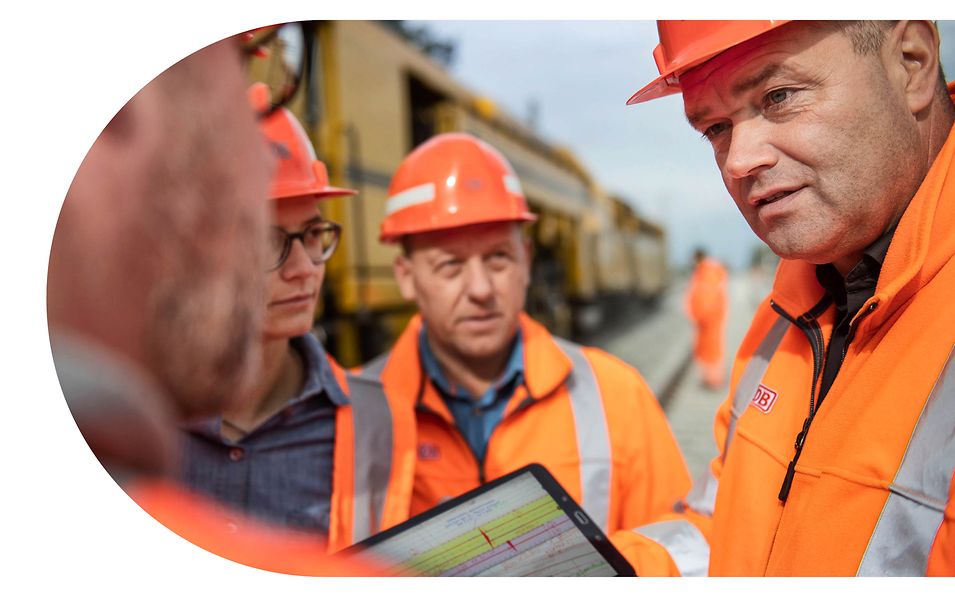 Vier Personen stehen in orangefarbener Schutzkleidung neben einer Gleisbaumaschine und besprechen den Plan auf einem Tablet. 