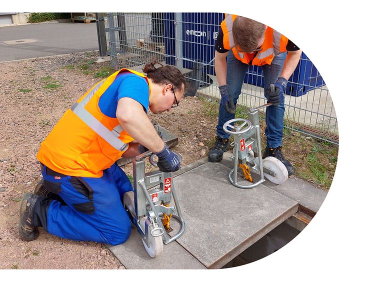 Zwei Männer in orangefarbener Schutzkleidung heben eine Steinplatte aus dem Boden. 