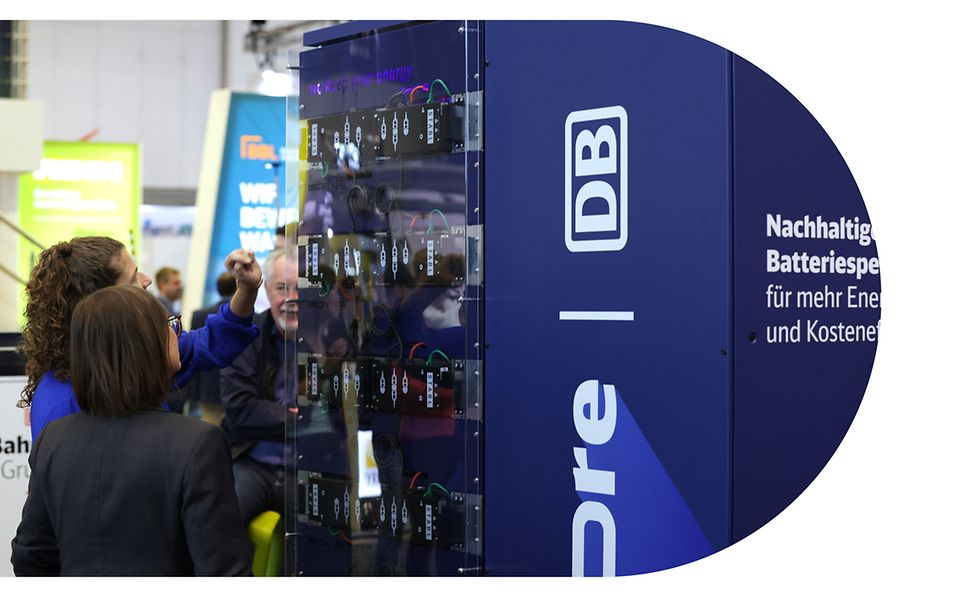 Zwei Personen stehen auf einer Messe seitlich von einem encore | DB Batteriespeicher und betrachten die Technologie.