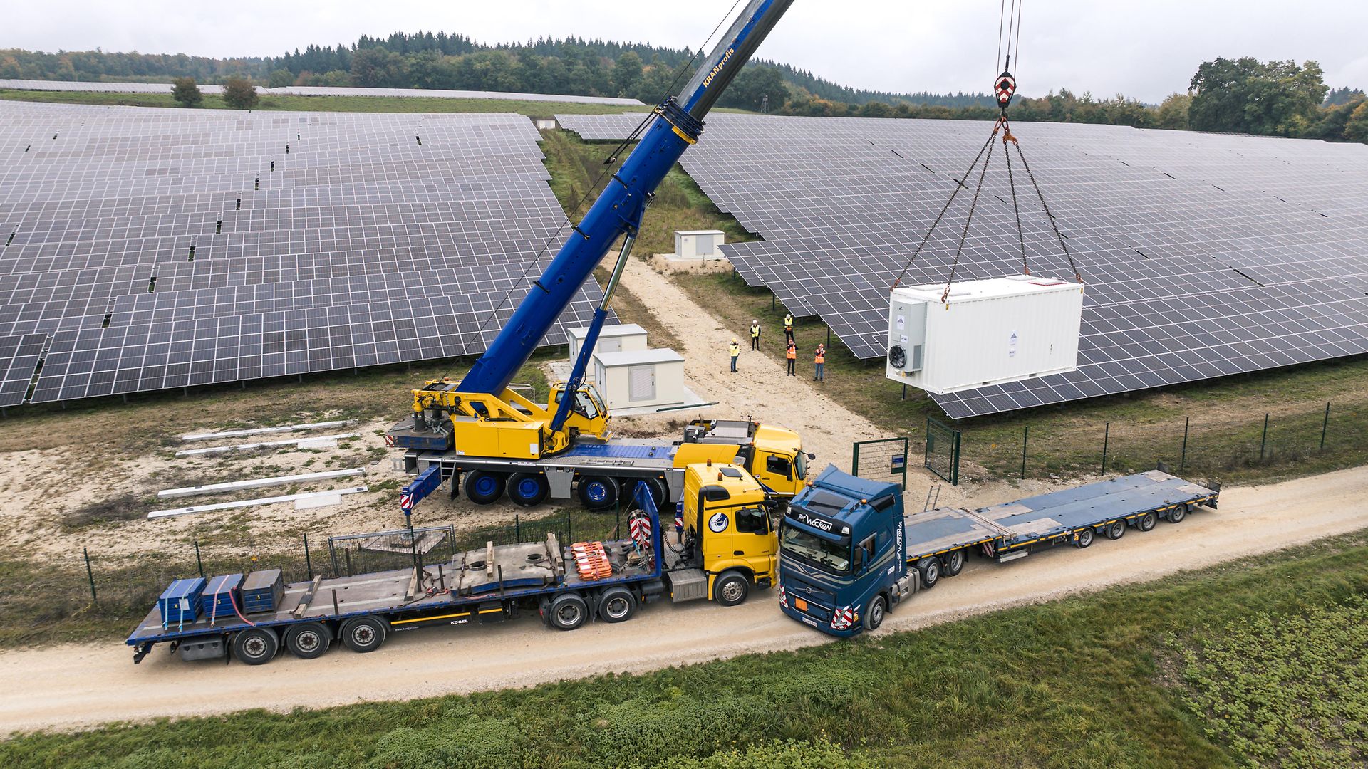 Anlieferung Batteriespeicher, 2 LKWs, 1 Kran, im Hintergrund Mitarbeitende und Solarpark
